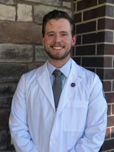 Zach Juaire standing in a white dental coat in front of a brick wall.